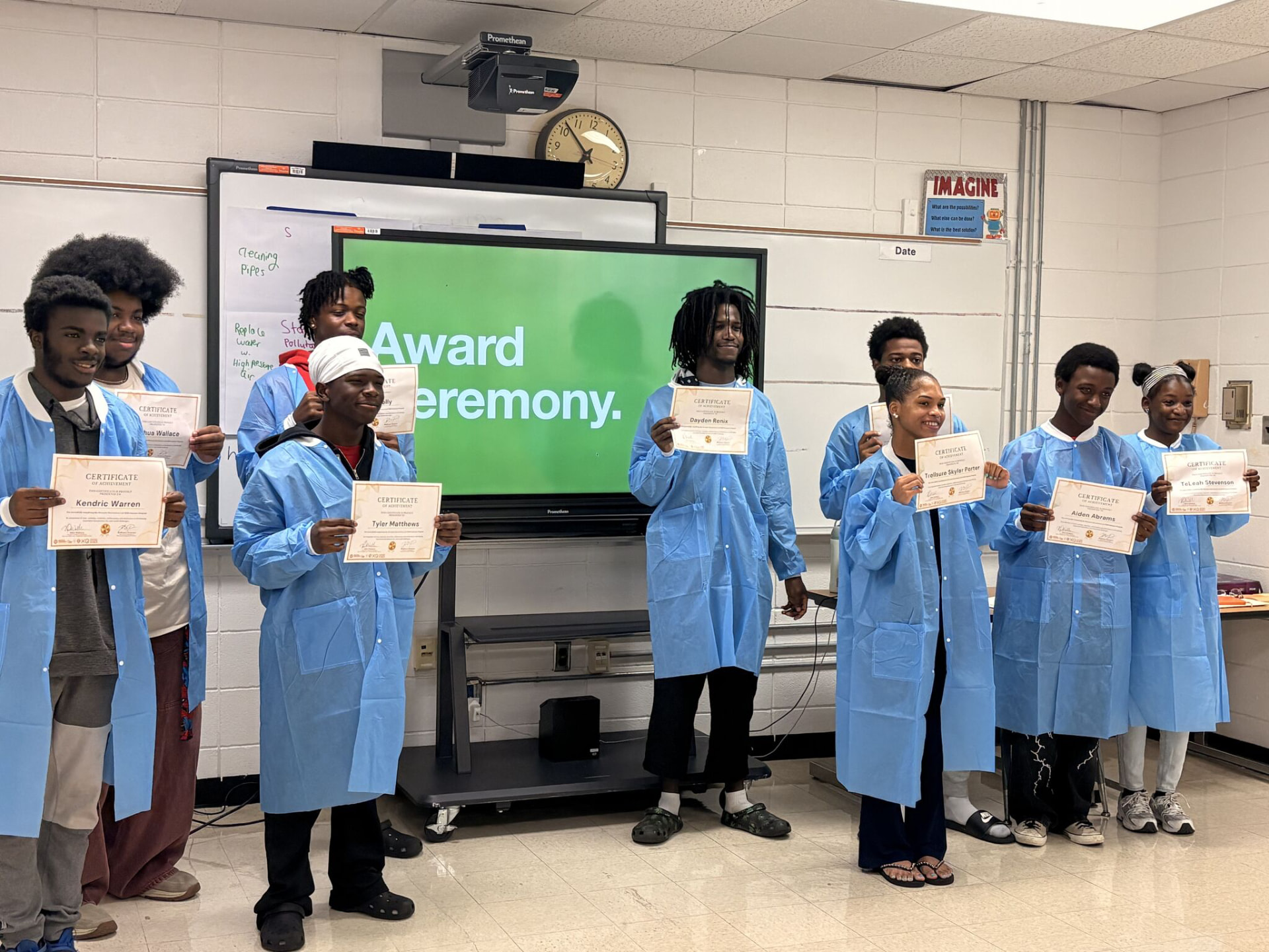 A group of young scientists in blue lab coats holding their certificates at an award ceremony